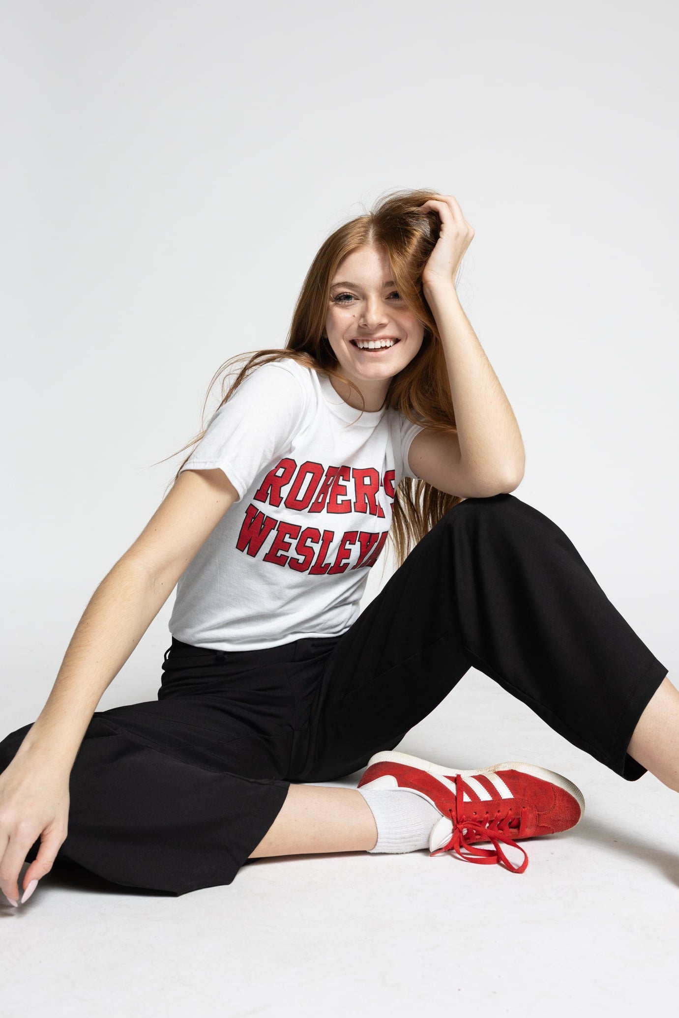 Person wearing a white Roberts Wesleyan t-shirt with red text, black pants, and red sneakers on a white background