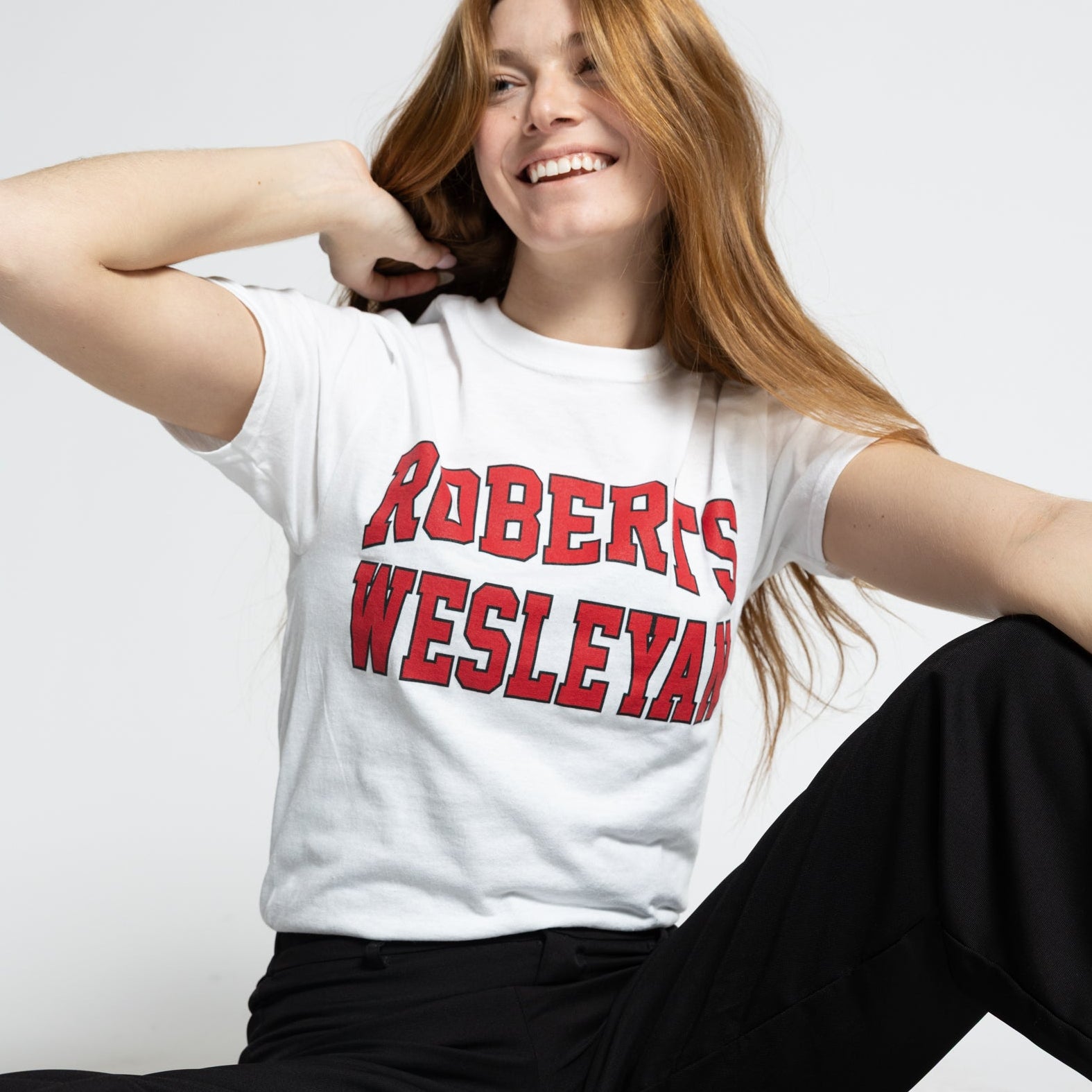 Person wearing a white Roberts Wesleyan t-shirt with red text, black pants, and red sneakers on a white background