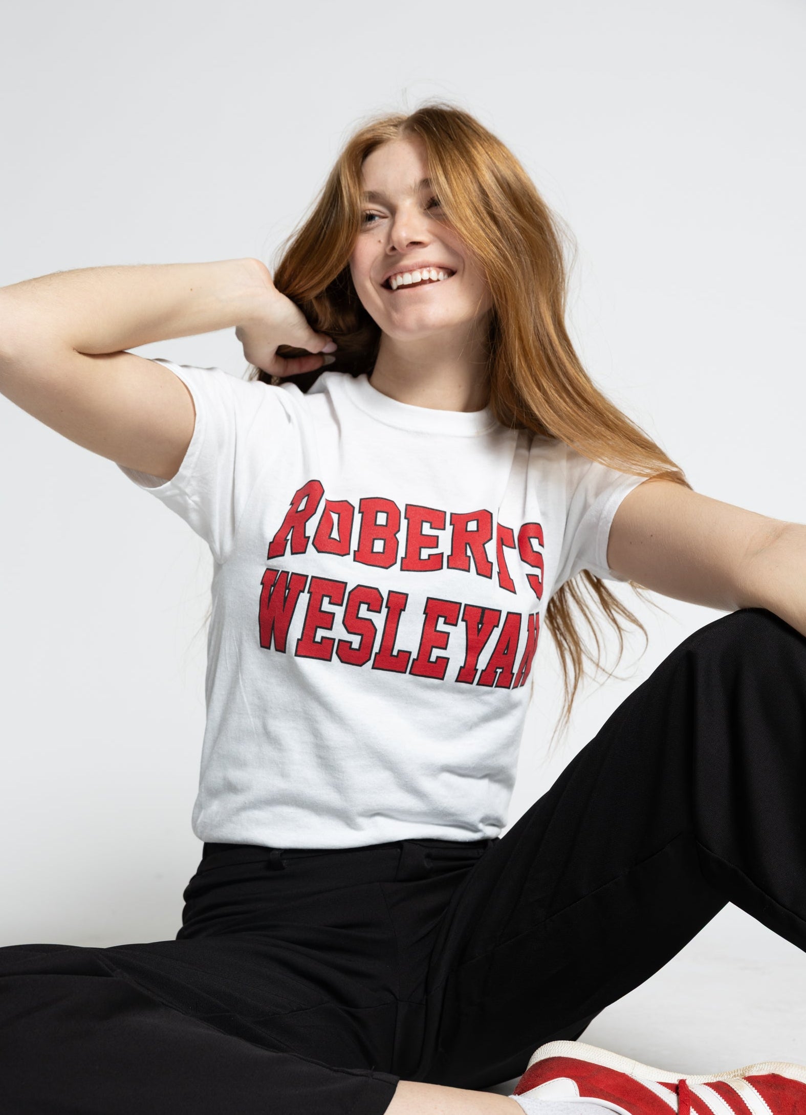 Person wearing a white Roberts Wesleyan t-shirt with red text, black pants, and red sneakers on a white background