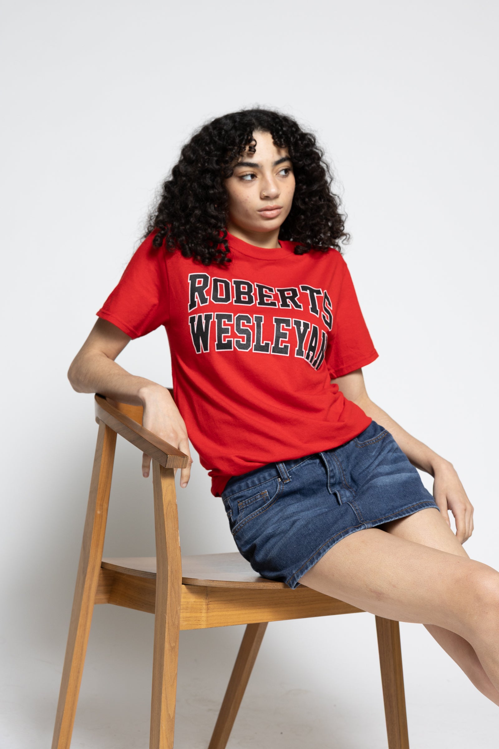 Person wearing a red 'Robert Wesleyan' t-shirt sitting on a wooden chair against a white background