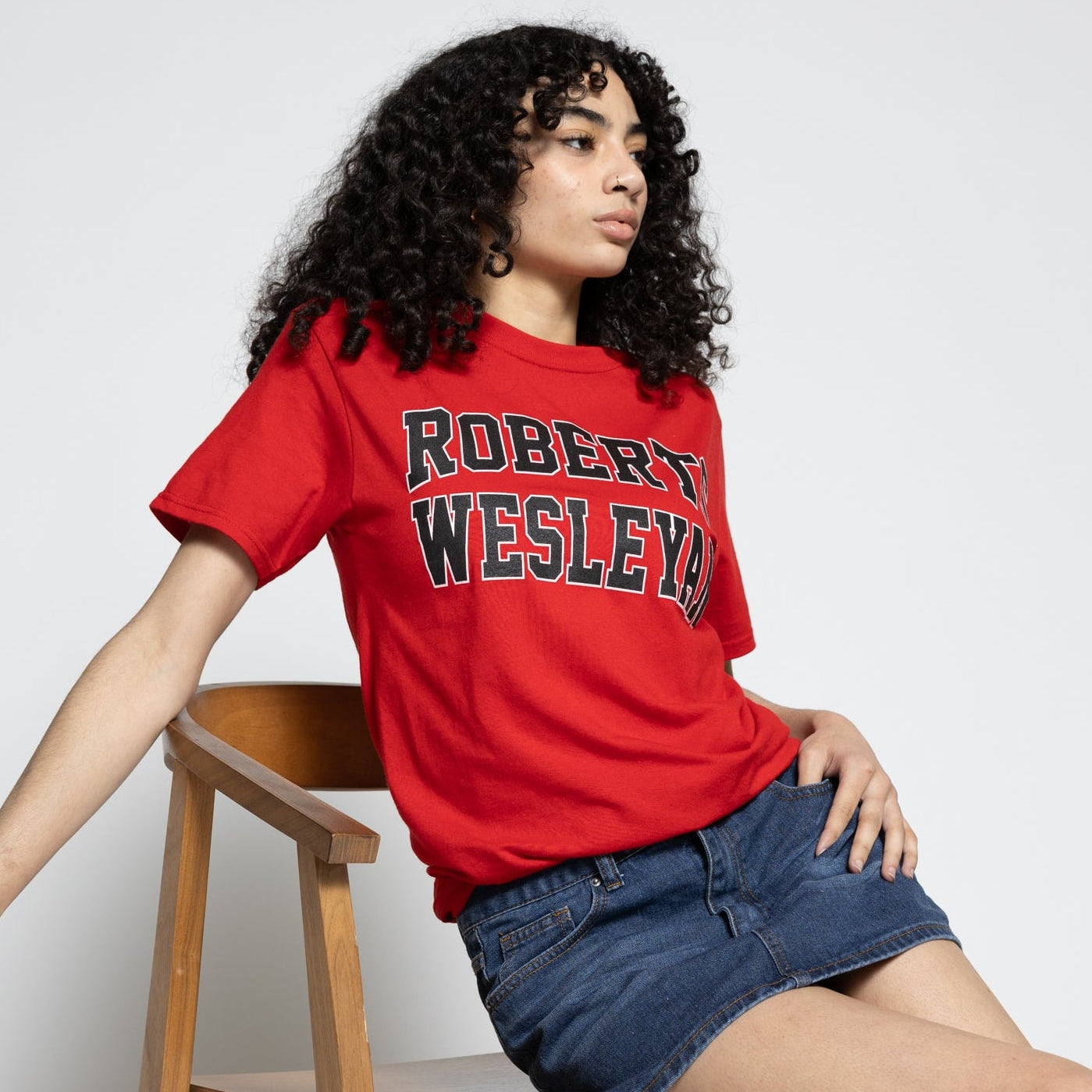 Person wearing a red 'Robert Wesleyan' t-shirt sitting on a wooden chair against a white background