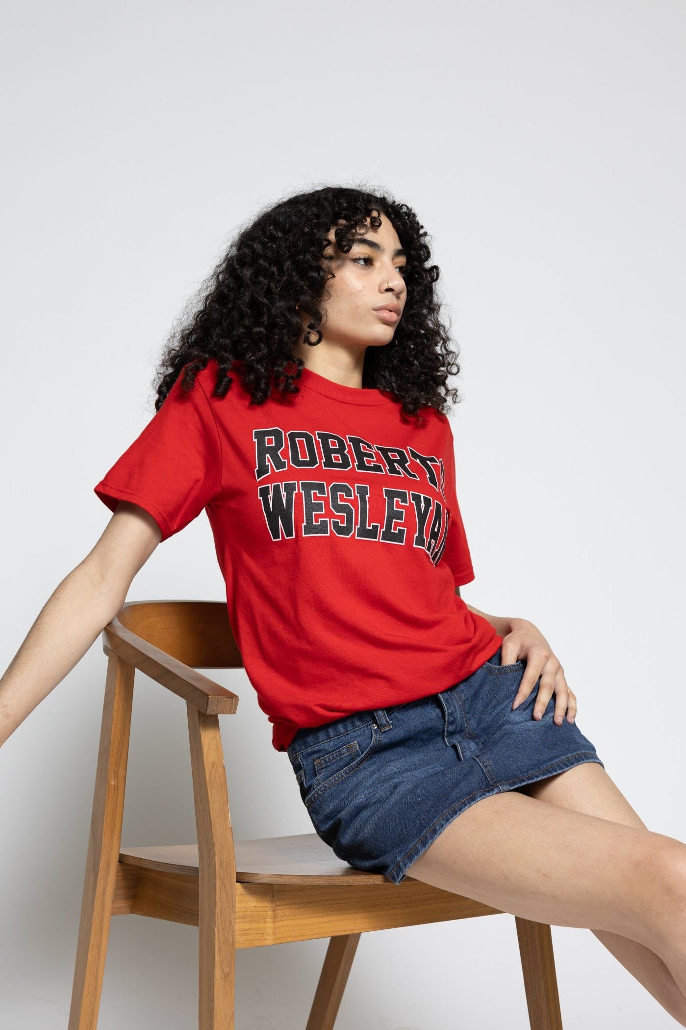 Person wearing a red 'Robert Wesleyan' t-shirt sitting on a wooden chair against a white background