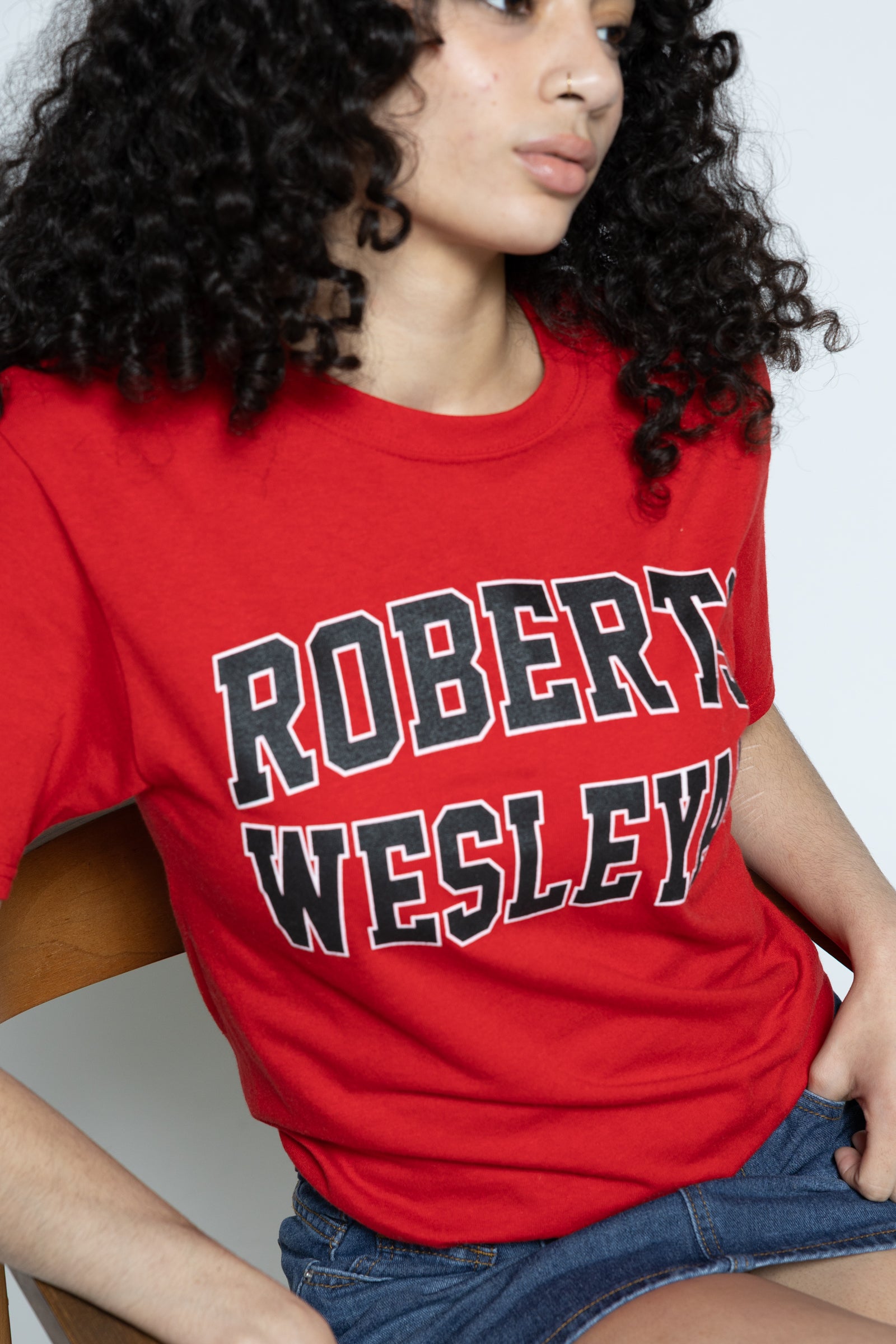 Person wearing a red 'Robert Wesleyan' t-shirt sitting on a wooden chair against a white background